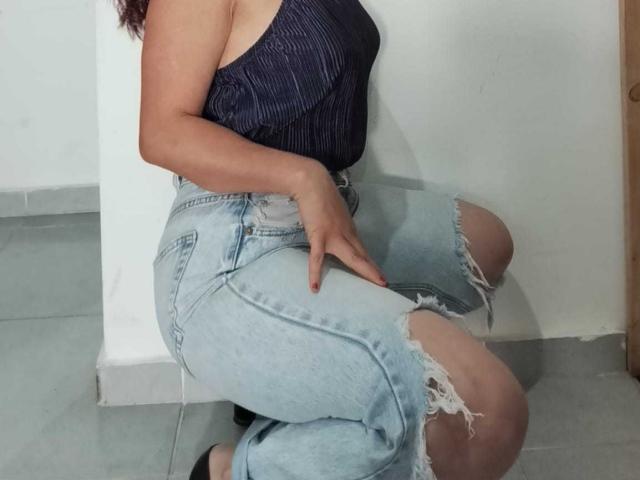 CamileParist, with medium brown skin, wearing a dark blue pinstripe top and ripped light blue jeans, sits on the floor, touching her knee, showing her red-painted nails.