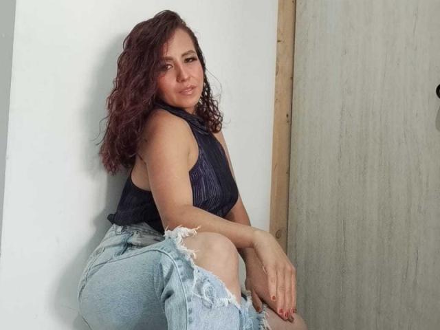CamileParist has curly red-brown hair, a dark sleeveless top, and ripped blue jeans, sitting with hands on knees, showing her ass and a slight smile.