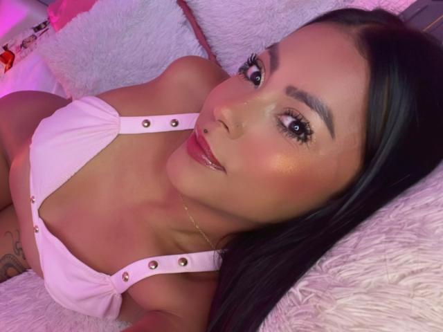 Amberv has dark hair, a nose ring, and is wearing a pink top with gold buttons, showing her tits and a tattoo on her left side. She's lying on a white pillow.