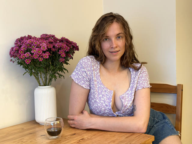 KenzieONEx with wavy brown hair, wearing a purple floral top with a deep cleavage, sits at a wooden table, arms crossed, next to a vase of pink flowers, and a glass.