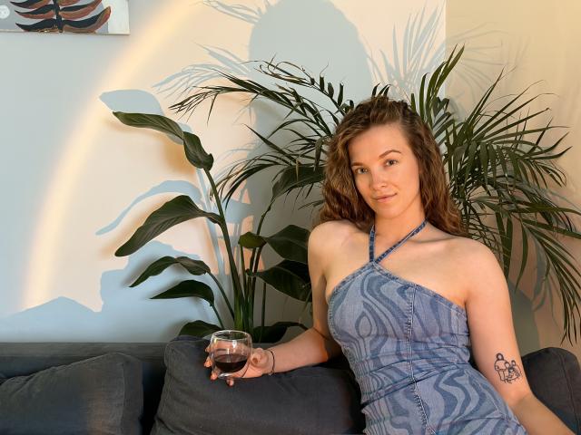KenzieONEx with wavy brown hair, wearing a blue patterned halter dress, holding a glass of dark wine, has a tattoo on her right arm, sitting on a gray couch.