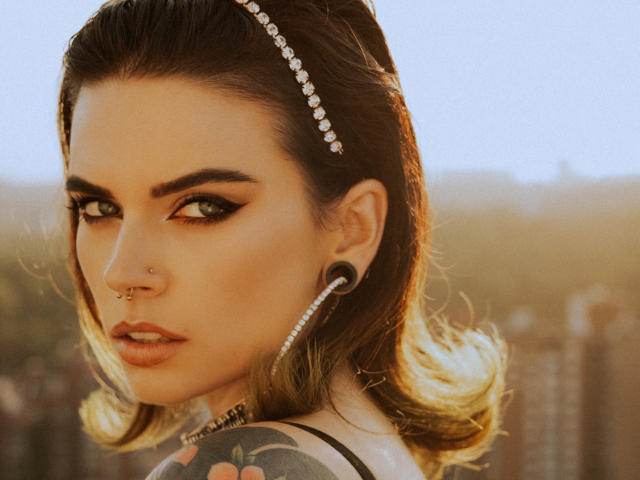SerenaMilss has dark brown hair with a diamond headband, black eyeliner, and large hoop earrings. She has a nose ring, tattoo on her shoulder, and is looking back.