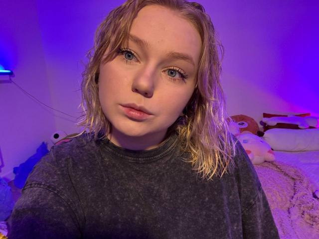 LimeCaty with wet blonde hair, blue eyes, and a serious expression, wearing a dark shirt, in a room with purple lighting. Her face is centered, with a bed behind.