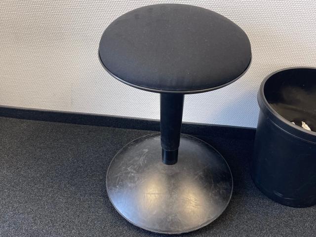 A black, round-topped stool with a matte finish and a thick, cylindrical base is placed on a dark carpet, next to a black cylindrical trash can.