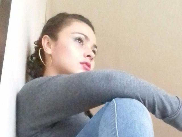 LusciousX, con labios rosados y pendientes de aro, está sentada con las rodillas levantadas, vistiendo una camiseta gris de manga larga y jeans azules, con su cabello oscuro y rizado recogido..
