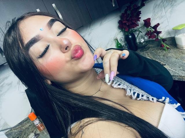 AnaShalimarr, lábios rosados e volumosos, olhos fechados, cabelo escuro, delineador azul, blush rosa, colar branco, manga preta, tocando o queixo, sutiã azul com detalhes brancos, unhas roxas, bancada da cozinha..