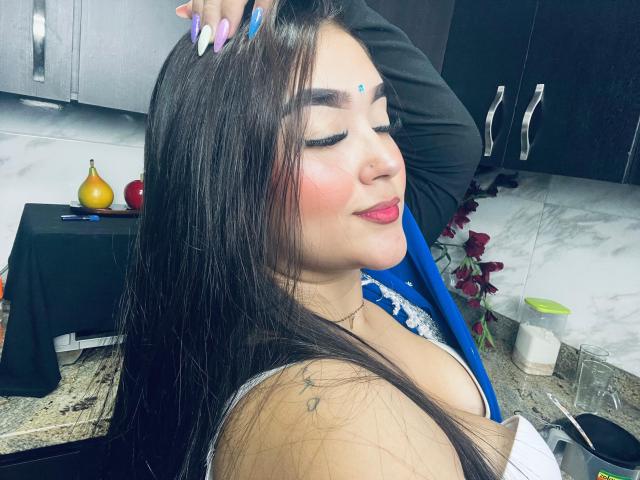 AnaShalimarr mit langem, schwarzem Haar, geschlossenen Augen, pinkem Lippenstift und einem blauen Punkt auf der Stirn, hat ihre Brüste leicht sichtbar in einer Küche, wo eine schwarze Hand sie berührt..