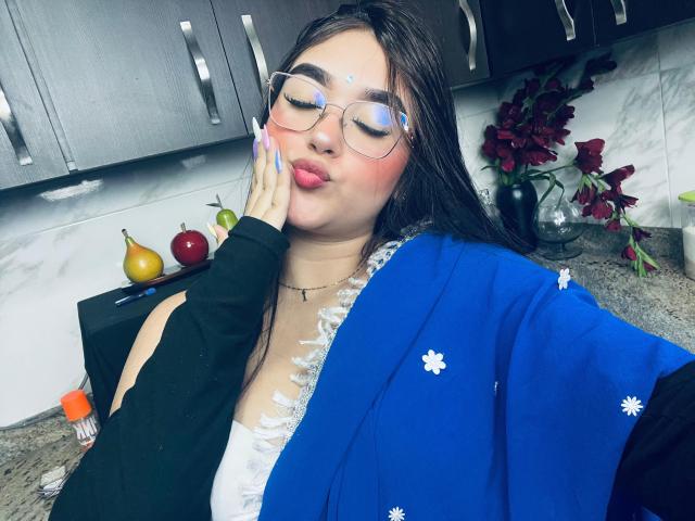 AnaShalimarr with glasses, pink lips, and blue shawl, touching her cheek, showing her tits in a white lace top, in a kitchen. Her long dark hair and fair skin.
