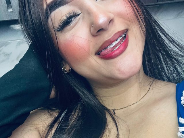 AnaShalimarr has long black hair, a nose piercing, red lipstick, and is sticking out her tongue with her teeth showing. She's wearing a gold necklace and has pink blush.