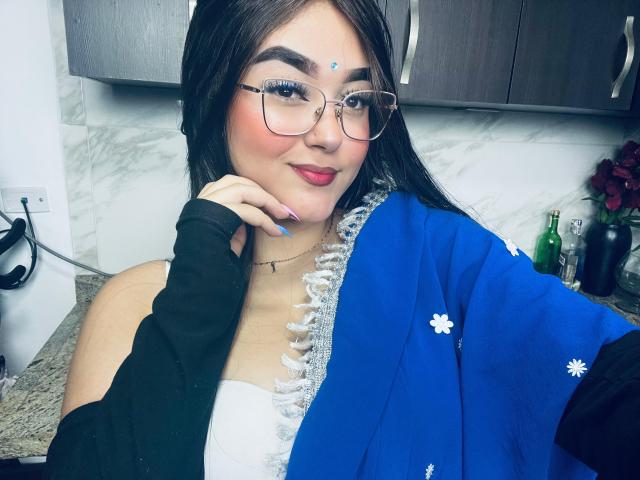 AnaShalimarr mit Brille, rotem Lippenstift, blauem Blumenbademantel, schwarzem Oberteil, weißem Kleid, blauen Nägeln, schwarzen Haaren, weißem Punkt auf der Stirn, lächelnd, Hand am Wangenknochen, Küchenhintergrund..