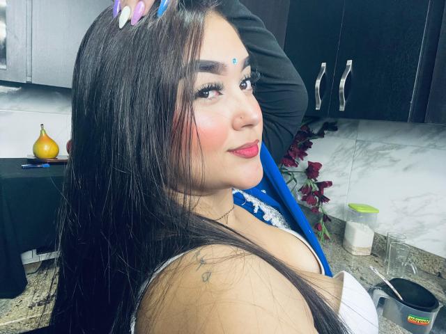 AnaShalimarr, com cabelos longos e escuros, um ponto azul na testa, batom rosa e uma blusa de renda azul, mostrando os seios, em uma cozinha, usando um sutiã de renda azul e branco..