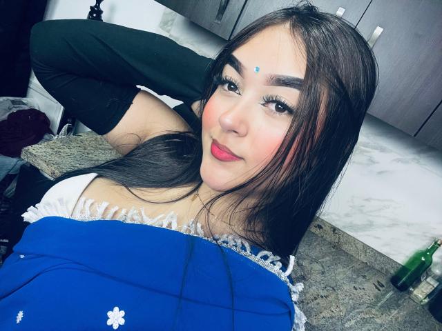 AnaShalimarr, con cabello castaño largo, ojos azules y un pequeño punto azul en la frente, acostada sobre una encimera de cocina, vestida con una blusa negra y un vestido azul, sonriendo..