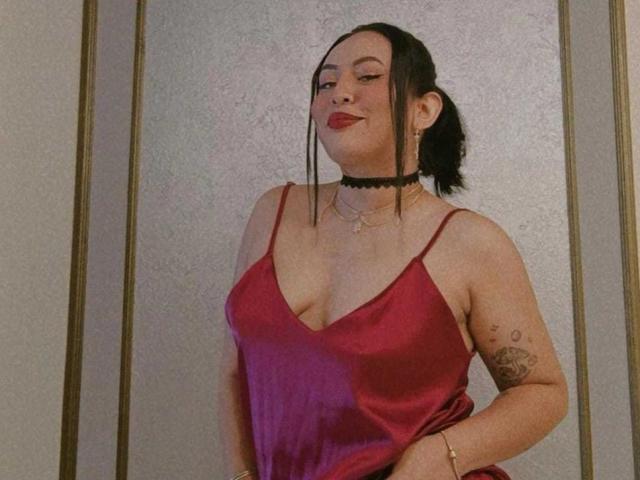 LiliethWolf in a red satin dress, black choker, and ponytail, showing her tits, tattooed arm, and smiling with red lipstick, standing against a textured wall.