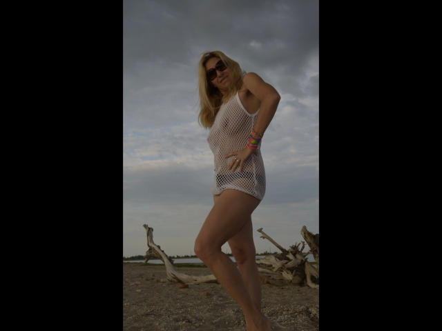FastOrgazm4U-hot wearing a white mesh dress, showing her tits and ass, standing with one leg bent, blonde hair, sunglasses, colorful bracelet, against a cloudy sky.