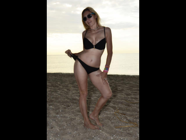 FastOrgazm4U-hot standing on a beach, wearing a black bra and panties, pulling down her panties, with sunglasses on, and colorful bracelets, at sunset.