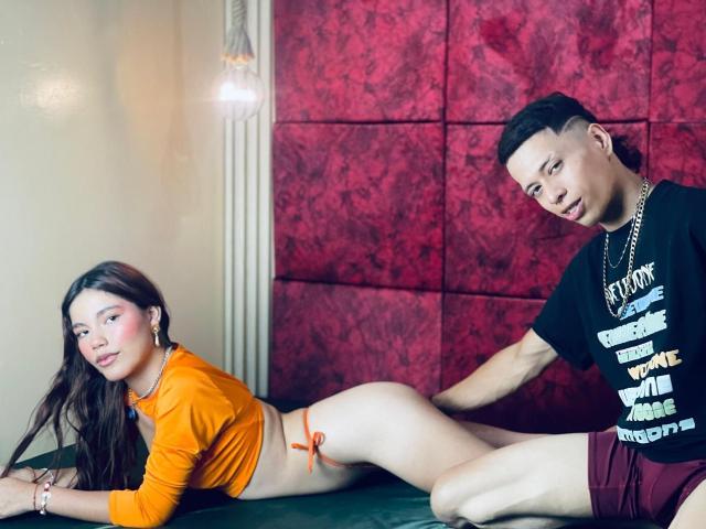 LiaDani with long brown hair, orange top, and no bottom is on her knees, facing left, while a guy in a black shirt and red shorts touches her ass.