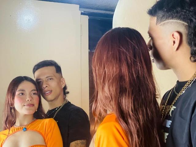 LiaDani with red hair, orange top, showing her tits, stands next to a guy with a mohawk, black shirt, and gold chain, in a mirror selfie.