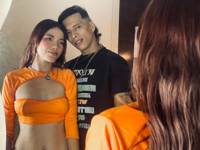 LiaDani with long brown hair in an orange crop top showing her tits, standing next to a boy with a mohawk in a black shirt, both looking at a mirror.
