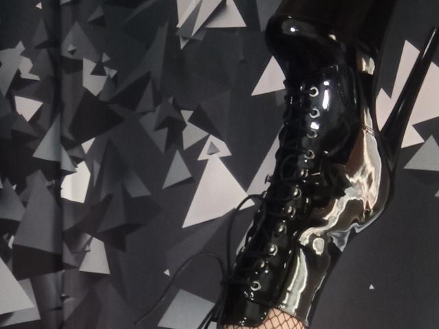 MonicaMiss's shiny black leather boot with laces and fishnet, showing a white splash on the side, angled to the right. Boot dominates the frame.