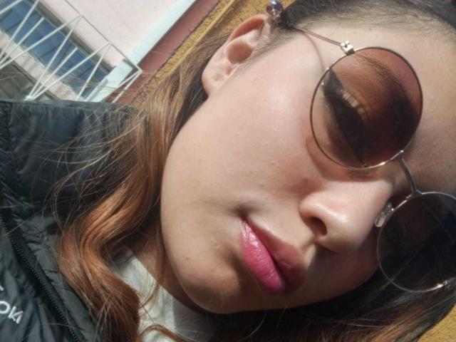 DominicTheOne has tan skin, brown eyes, and glossy pink lips, wearing round sunglasses with reflective brown lenses, and a black jacket. She has brown hair with subtle highlights.