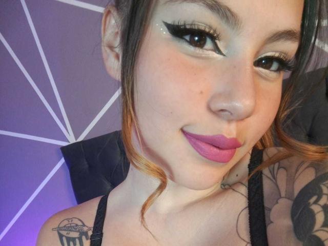 DominicTheOne has fair skin, black eyeliner, purple lipstick, and a small nose ring. She's smiling, showing off tattoos on her shoulder. Her brown hair is partially tied up.