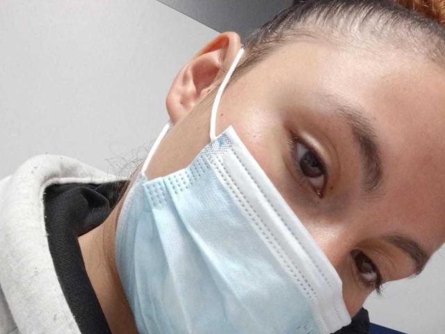 DominicTheOne's face with a blue surgical mask covering the lower half, brown eyes looking slightly upwards, black shirt, white hoodie, and brown hair pulled back.