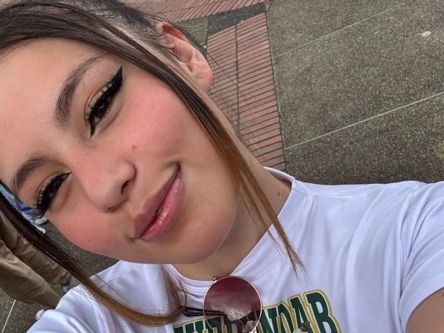 DominicTheOne has light brown skin, black eyeliner, and a small smile. She's wearing a white shirt with her brown hair in a ponytail, and has sunglasses on her chest.