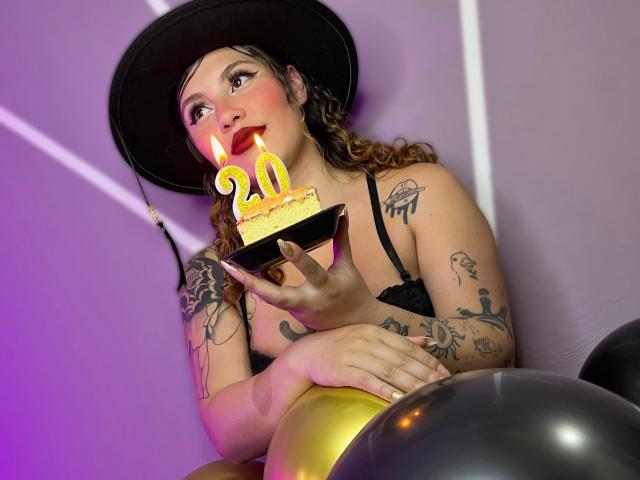 DominicTheOne with curly brown hair, black hat, and tattoos, holding a cake with a yellow "20" candle, pink lipstick, black bra, and gold balloon in the foreground.