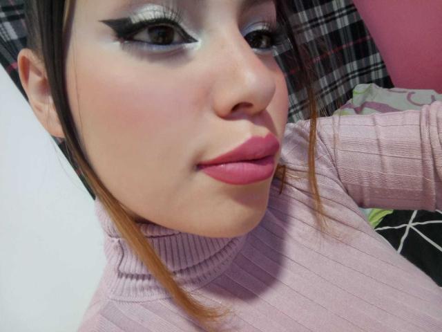 DominicTheOne has light skin, pink lipstick, and a pink turtleneck. She's got dramatic eyeliner and brown eyes, with blonde hair pulled back. Her lips are slightly pouty.