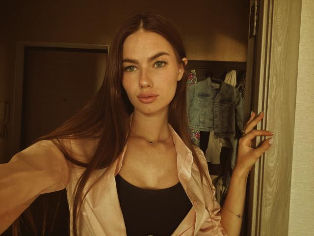 LadyJRouses with long brown hair, green eyes, and pouty lips, wearing a pink satin shirt over a black top, standing in a doorway, hand on curtain.