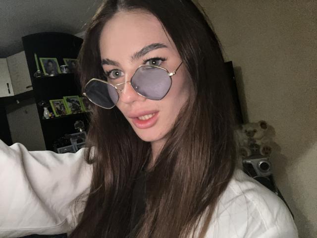 LadyJRouses with long brown hair, wearing round glasses, white shirt, and pink lipstick, showing her small tits and slightly open mouth in a selfie.