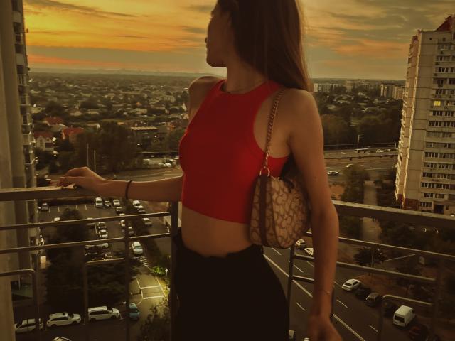 LadyJRouses with red crop top, black high-waisted pants, and brown handbag, standing on balcony at sunset, holding railing, city skyline and cars below, long brown hair, side profile.