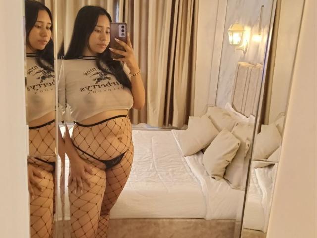Karollteyes with long black hair, wearing a sheer white top and black fishnet pants, showing her ass and tits in a mirror selfie, standing in a bedroom.