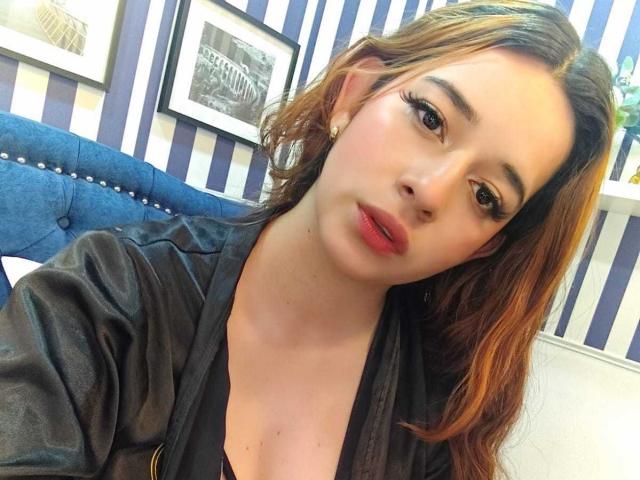 DaniVelasquez with pouty red lips, brown wavy hair, black eyeliner, and pearl earrings, wearing a black shirt, sitting on a blue upholstered chair.