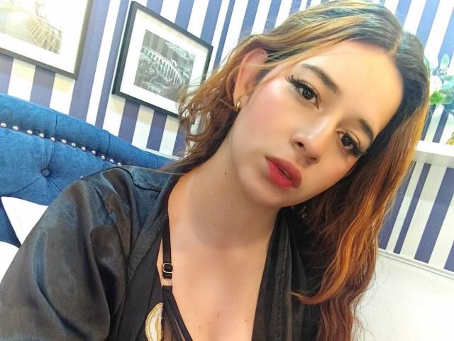 DaniVelasquez with pouty red lips, long brown hair, black shirt, and eyeliner, looking at camera, showing her boobs in a low-cut top. Striped wall and blue chair in background.