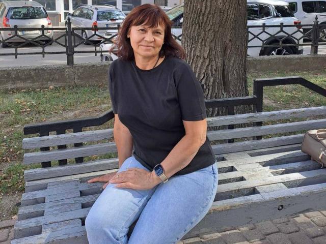 Ingrid Wels, com cabelo ruivo, camiseta preta e calça jeans azul, sentada em um banco de madeira cinza, usando um relógio azul, olhando ligeiramente para a direita..