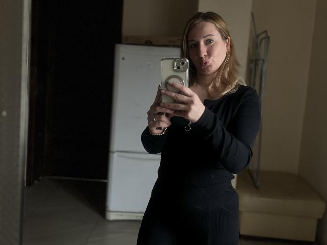 MilaqwRouse with blonde hair, pouty lips, and medium tits, wearing a black long-sleeve dress, taking a selfie in a dimly lit room, holding a phone with a ring.
