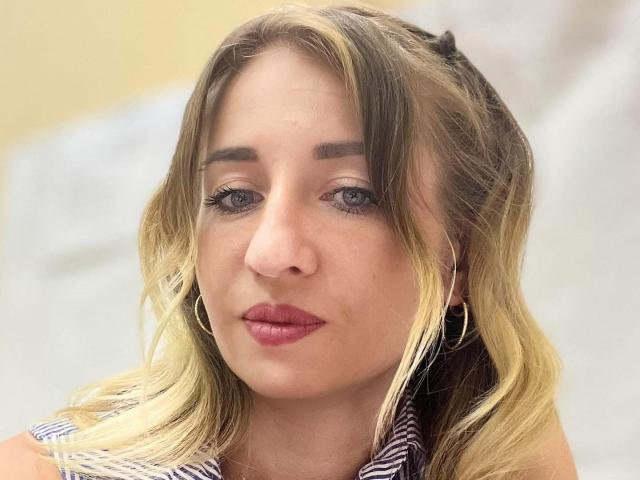 MilaqwRouse has blonde hair, blue eyes, and red lipstick. She's wearing a striped shirt with hoop earrings. Her face is slightly pouty with a subtle smile.