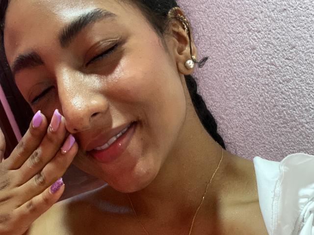 NatalyBlonder has her eyes shut, smiling with a hand touching her cheek. She has tan skin, purple nails, and is wearing a pearl earring and a white top.
