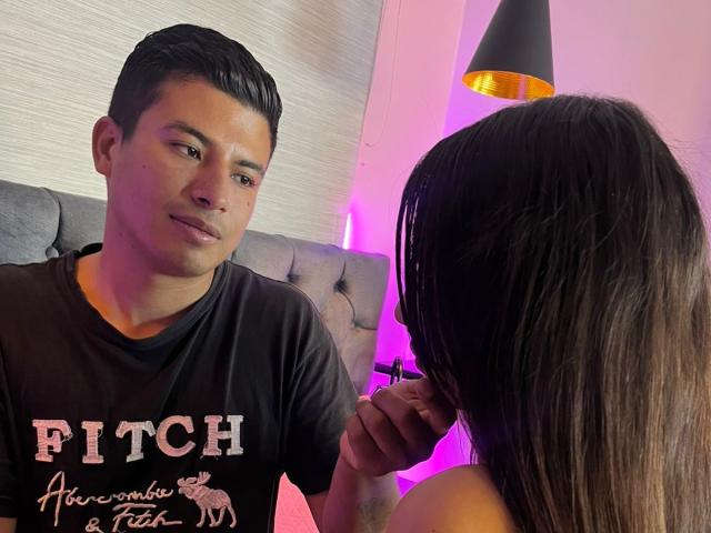 DangerAllie with short black hair, slight smile, black t-shirt, sitting, looking at another person with long brown hair, DangerAllie's hand in hair, pink and yellow lighting, black lamp.