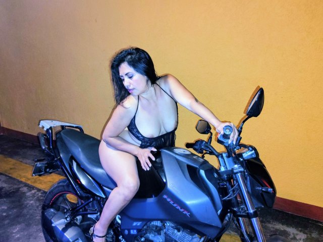 NatashaMonttero with big tits sitting on a black motorcycle, wearing a black lace dress, hands on the handlebars, looking to the side, in a dimly lit, yellow-walled room.