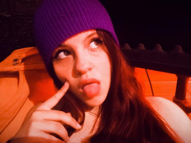 SophieHill with a purple beanie, sticking her tongue out, and pointing at her nose with her finger, has brown hair and a nose ring, in a warm, orange-lit setting.
