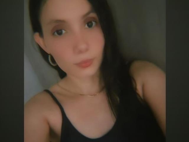 HallieMils with dark hair, wearing a black tank top, small tits, light skin, wearing gold hoop earrings and a thin necklace, in a slightly blurry selfie.