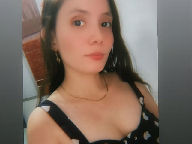 HallieMils has light skin, brown hair, and is wearing a black dress with white hearts, showing her small tits and a thin gold necklace, looking slightly to the left.