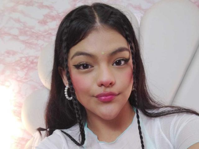 NikyBonnie has dark braided hair, pink lipstick, and eyeliner. She's wearing a white top with pearl earrings. Her ass is not visible. Her face is cute and she's smiling.