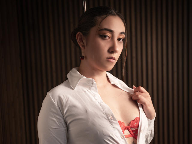SwettieSammy with light skin and brown hair, wearing a white shirt open to reveal red lace bra, holding shirt with right hand, looking down with subtle smile.