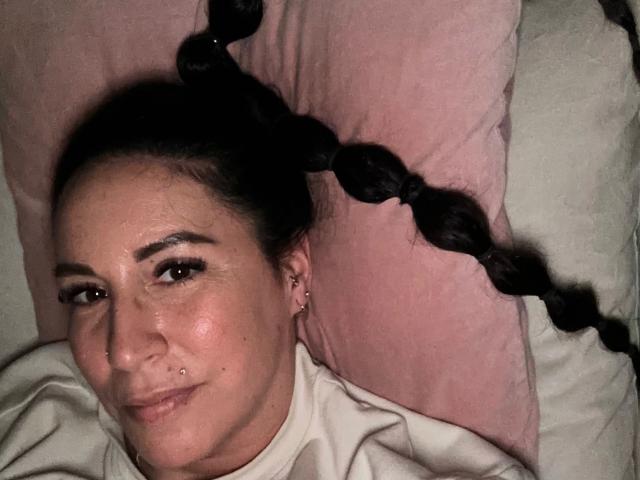 Greekmadame1-hot with long black braids lying on a pink pillow, wearing a white shirt, has a nose ring, and a subtle smile, showing a slight blush on her face.