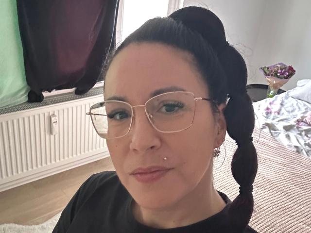 Greekmadame1-hot with black hair in a ponytail, wearing glasses, black shirt, nose piercing, and earrings, smiling in a room with clothes and a radiator.