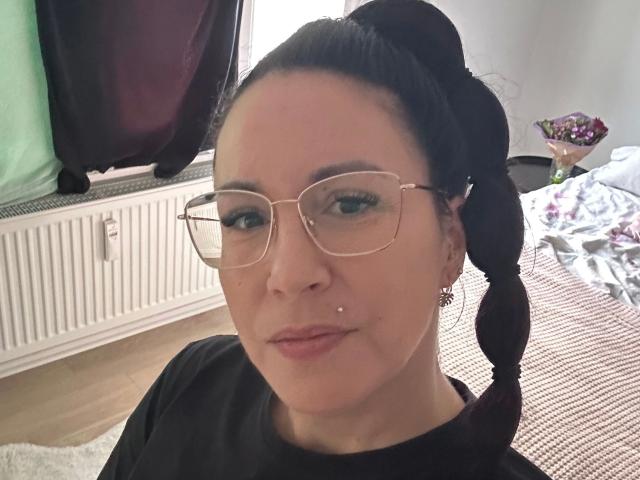 Greekmadame1-hot with glasses, black hair in a ponytail, black shirt, earrings, nose piercing, and subtle makeup, smiling at the camera.