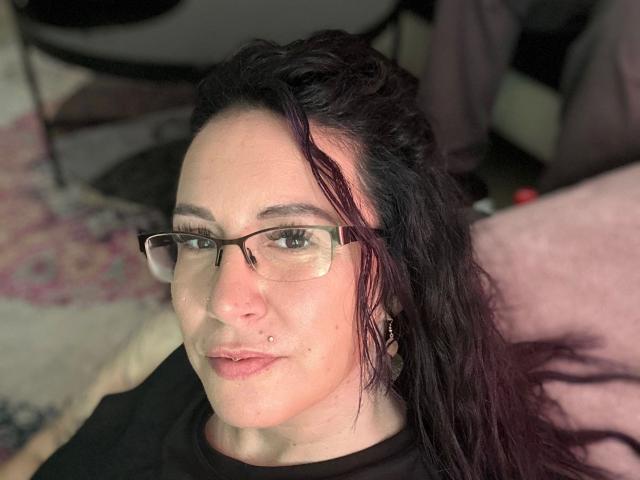 Greekmadame1-hot with dark wavy hair, glasses, nose piercing, and black shirt, showing a slightly pouty face. Her tits are small and she has a cute nose.